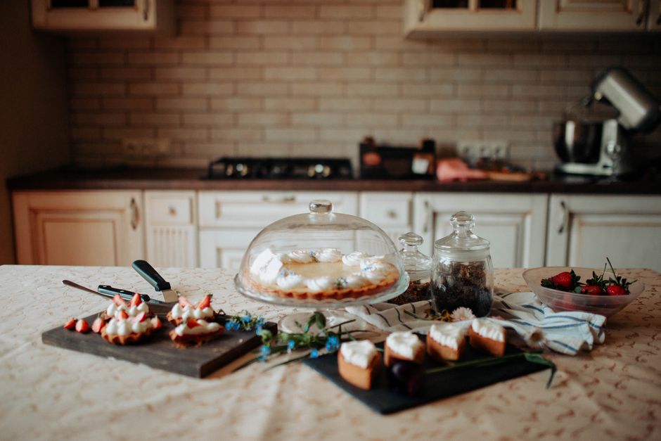 Imagem de uma mesa de cozinha decorada com comida caseira, incluindo bolo sob uma cobertura de vidro e frutas, transmitindo uma atmosfera acolhedora e rústica.