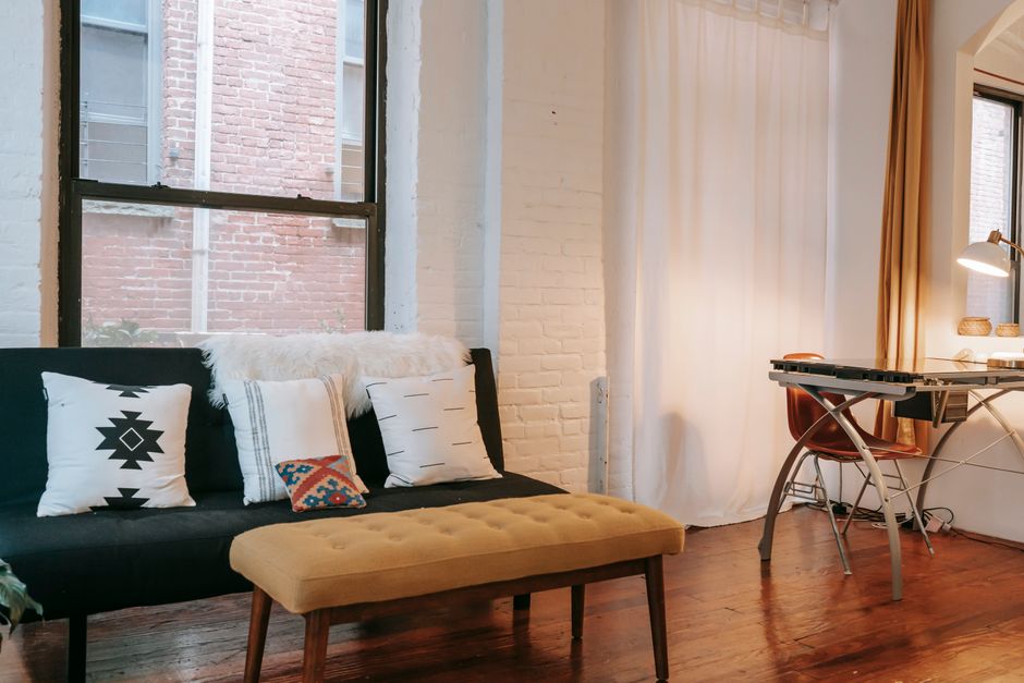 Imagem de um espaço de trabalho confortável em um ambiente residencial moderno, com sofá, almofadas e uma mesa de trabalho iluminada pela luz natural.