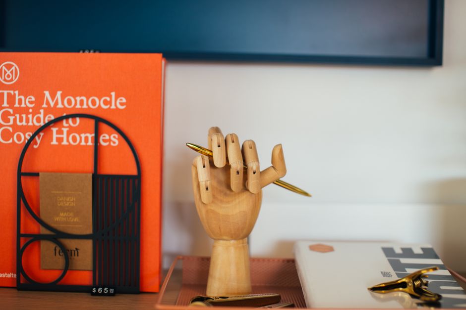 A imagem mostra uma composição de objetos de escritório, incluindo uma mão de madeira segurando uma caneta dourada, com livros e materiais de papelaria ao redor, transmitindo um ambiente criativo e organizado de trabalho ou estudo.