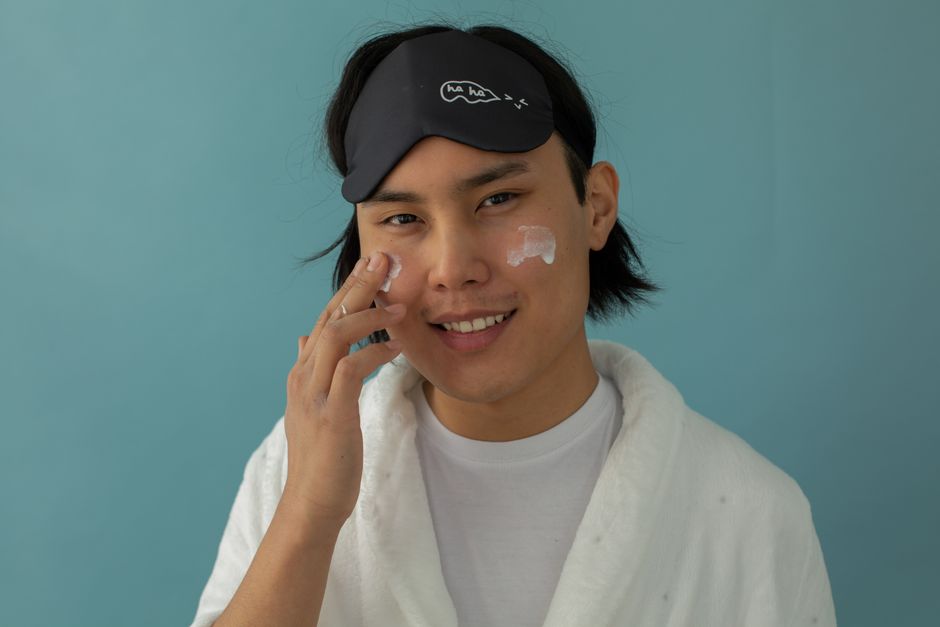 Pessoa sorridente usando máscara de sono e aplicando creme facial, promovendo cuidado com a pele e bem-estar.
