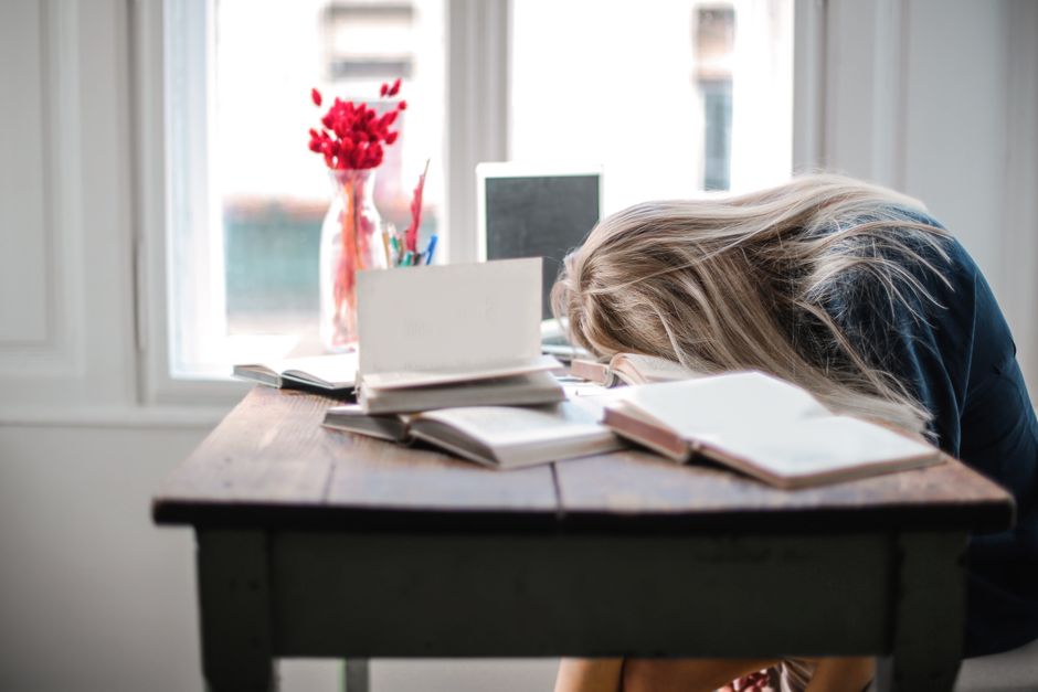 Pessoa com cabeça apoiada na mesa, rodeada de livros e materiais de estudo, refletindo cansaço ou esgotamento acadêmico ou profissional.