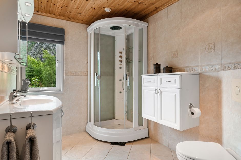 Banheiro moderno com ducha de cabine, bancada de pia, armário branco e janela com vista para a natureza.