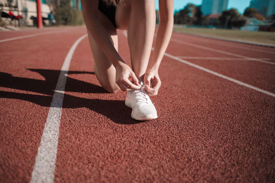 Mulher colocando cadarços em um tênis enquanto está em uma pista de corrida ao ar livre, simbolizando preparação e foco em atividades físicas ou esportes.