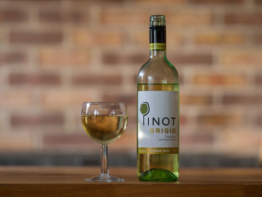 Imagem de uma garrafa de vinho branco ao lado de uma taça cheia, com fundo de parede de tijolos, sugerindo consumo de bebidas e momentos de lazer.