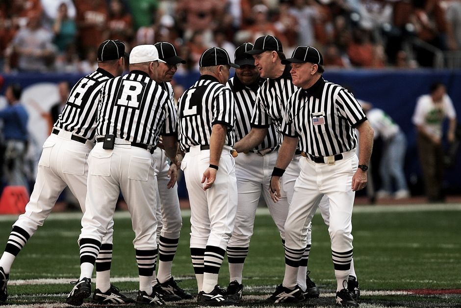 Grupo de árbitros de futebol em uniforme negociando ou discutindo durante uma partida na arena esportiva.