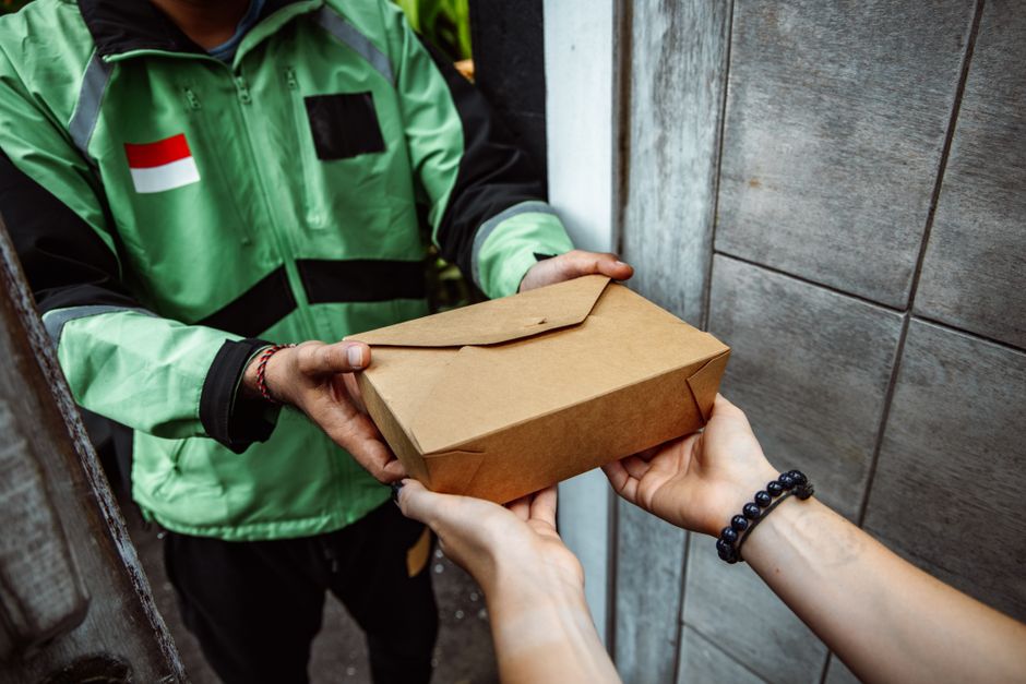 Pessoa entregando uma embalagem de comida para delivery em ambiente urbano ao ar livre.