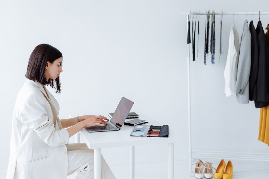 Mulher trabalhando no planejamento de coleção de moda em ambiente de consultoria de moda ou estilo, com roupas e acessórios ao fundo.