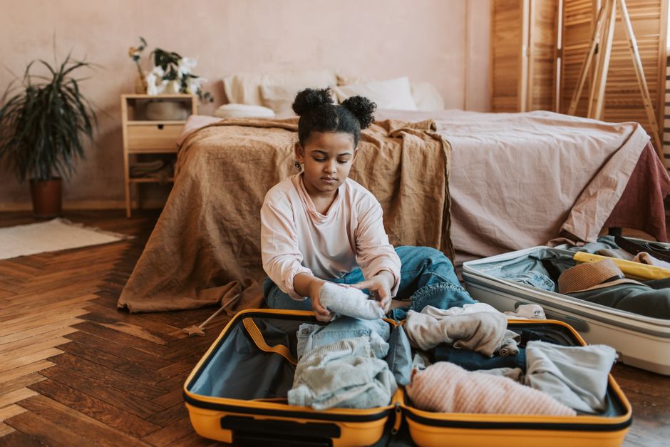Menina organizando roupa dentro de uma mala no quarto, sugerindo preparação para viagem ou mudança.
