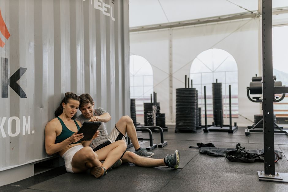 Dois jovens em uma academia de musculação descansando após o treino, compartilhando um momento de descontração com um dispositivo móvel.