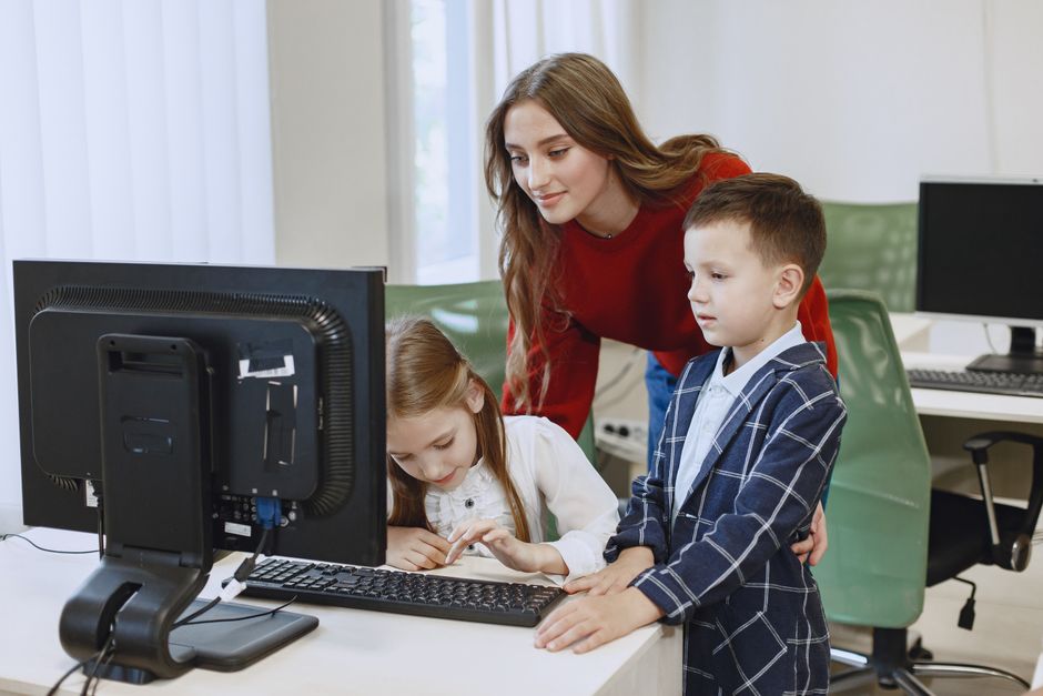 Uma mulher ajudando duas crianças a utilizarem um computador em um ambiente de aprendizado, refletindo colaboração e desenvolvimento infantil com tecnologia.