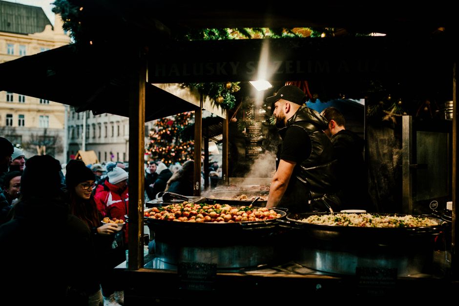 Homem preparando comida em uma barraca de rua durante um evento natalino ao ar livre, com várias pessoas ao redor apreciando o festival.