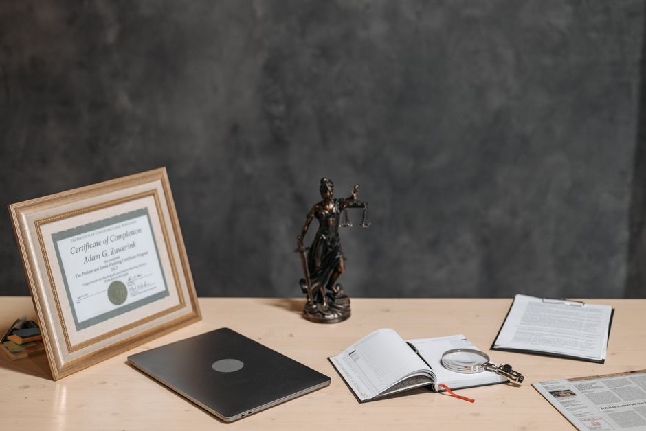 Imagem de uma mesa de trabalho com itens relacionados à área jurídica e educação, incluindo diploma, livro, balança da justiça e documentos.