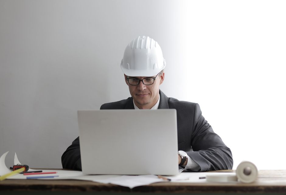 Homem de terno usando capacete de obra enquanto trabalha em um laptop, simbolizando gestão de projetos ou planejamento de construção.