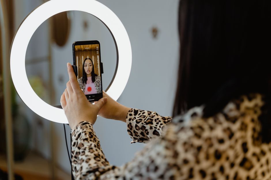 Pessoa tirando uma selfie diante de um espelho com luzes de ring light, evidenciando a produção de conteúdo ou maquiagem para blog ou redes sociais.