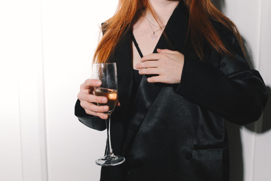Mulher com cabelo vermelho usando blazer preto segurando uma taça de vinho, simbolizando celebração ou eventos sociais.