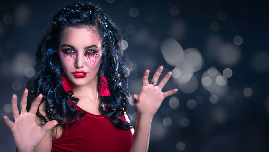 Mulher com maquiagem dramática e cabelo cacheado, usando roupa vermelha, em um fundo escuro com bokeh de luzes, evocando mistério ou expressão artística.