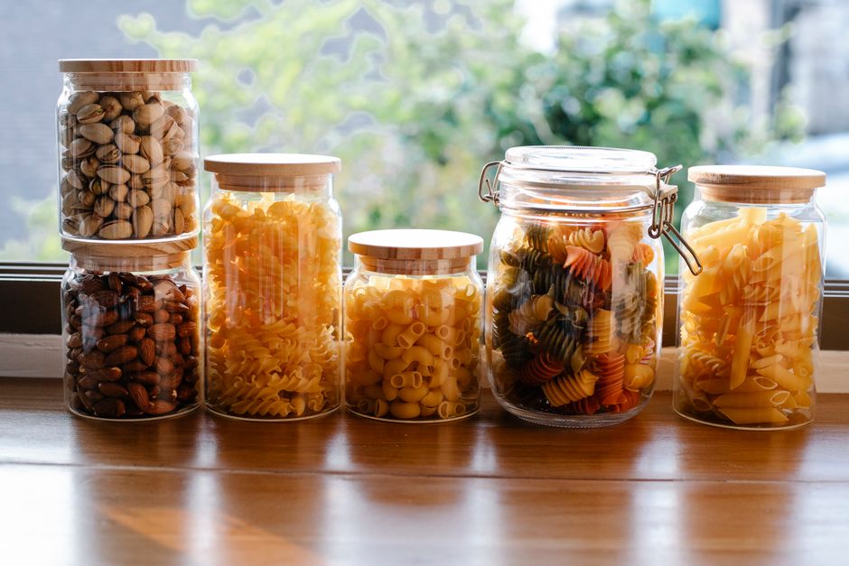 Imagem de potes de vidros recheados com diferentes tipos de massas coloridas, dispostos sobre uma mesa próxima a uma janela com vista para a natureza, representando organização na cozinha e preparo de refeições.