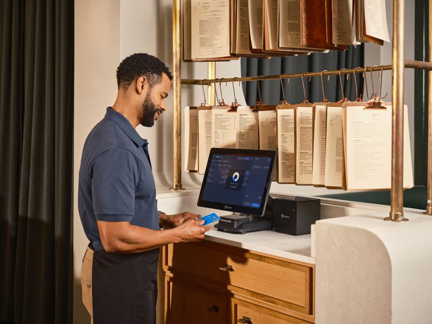 Homem usando um sistema de pagamento digital em um ambiente que parece ser uma cafeteria ou restaurante, com livros de receitas ou menus suspensos ao fundo.