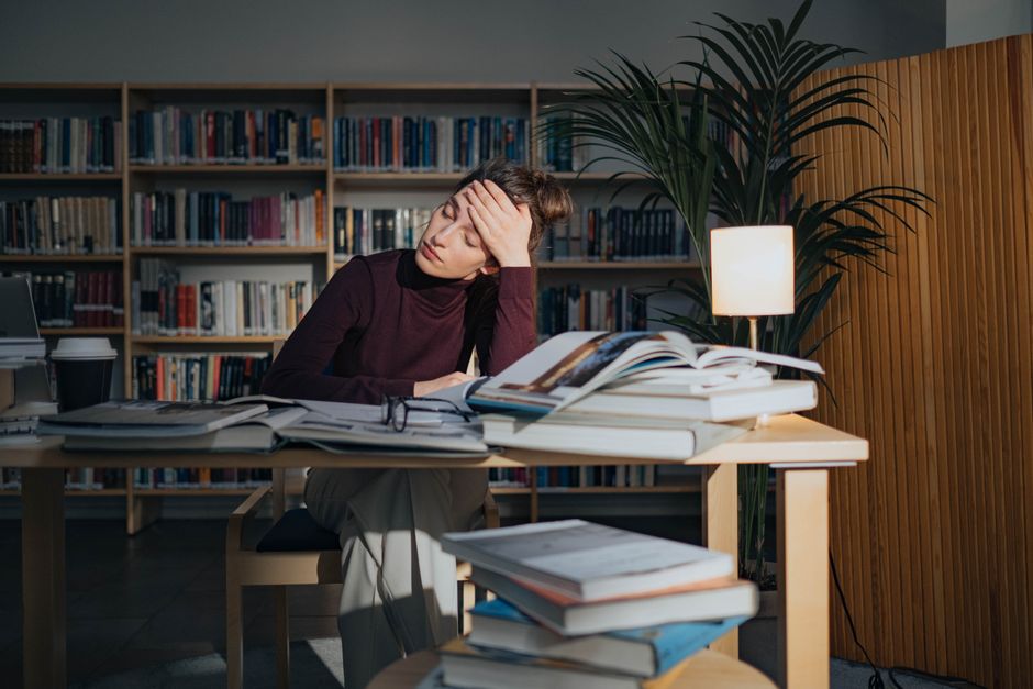 Pessoa estressada ou cansada, rodeada de livros e documentos, em um ambiente de estudo ou trabalho, refletindo sobre suas tarefas difíceis.