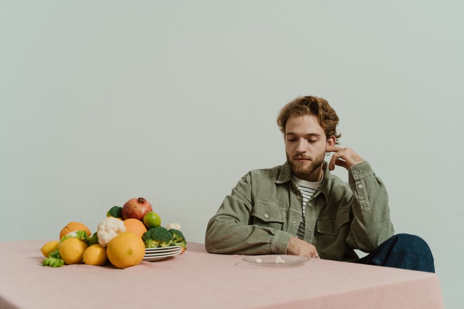 Homem pensativo sentado à mesa ao lado de uma variedade de frutas e legumes, indicando reflexão sobre alimentação saudável ou dieta.