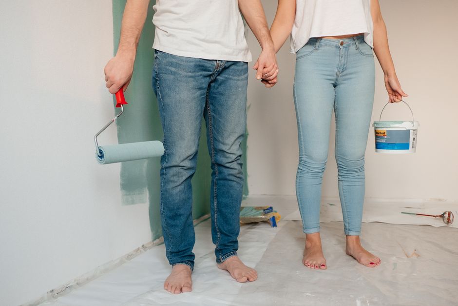 Imagem de um casal de mãos dadas enquanto pinta uma parede durante uma reforma residencial, simbolizando parceria e mudança em ambientes internos.