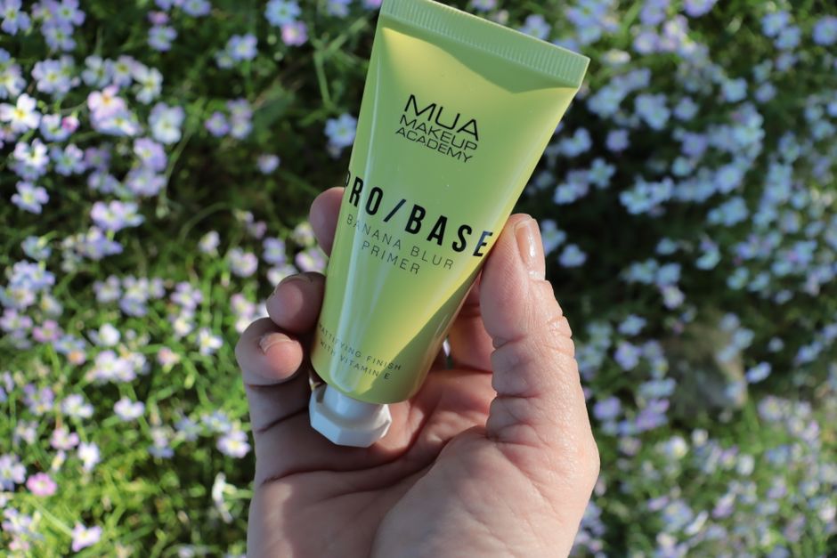 Pessoa segurando um tubo de primer de maquiagem com fundo de flores roxas e verdes, destacando produtos de beleza natural.