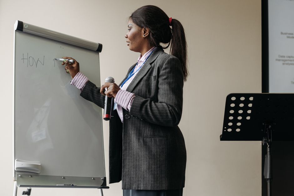 Mulher apresentando em um ambiente corporativo, utilizando um quadro branco para explicar um tema de negócios ou treinamento.