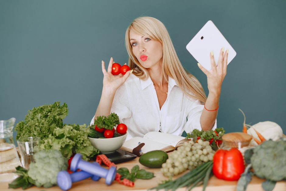 Mulher fazendo selfie em meio a alimentos saudáveis e utensílios de cozinha, transmitindo uma mensagem sobre alimentação e bem-estar.