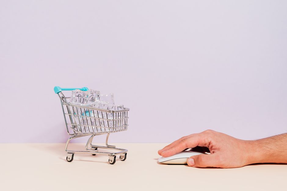 Imagem que representa comércio eletrônico ou compras online, com um carrinho de supermercado em miniatura cheio de produtos e uma mão usando um mouse de computador.