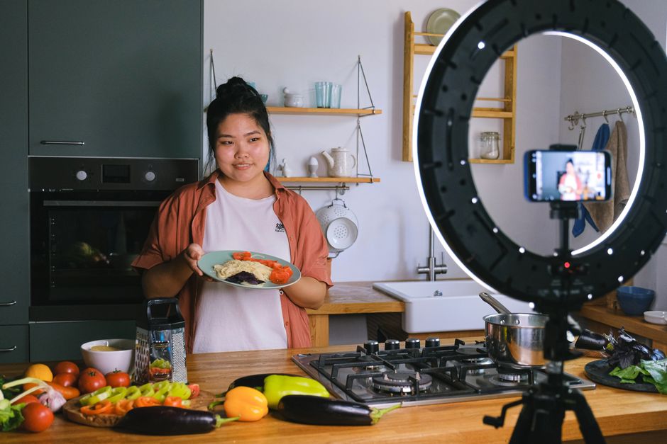 Jovem preparando e filmando um prato de comida em uma cozinha moderna, com iluminação de ring light para vídeos de receitas para blog ou canal de culinária.