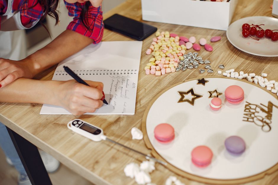 Pessoa anotando planos de celebração ao lado de doces e guloseimas decorativas em uma mesa de festa.