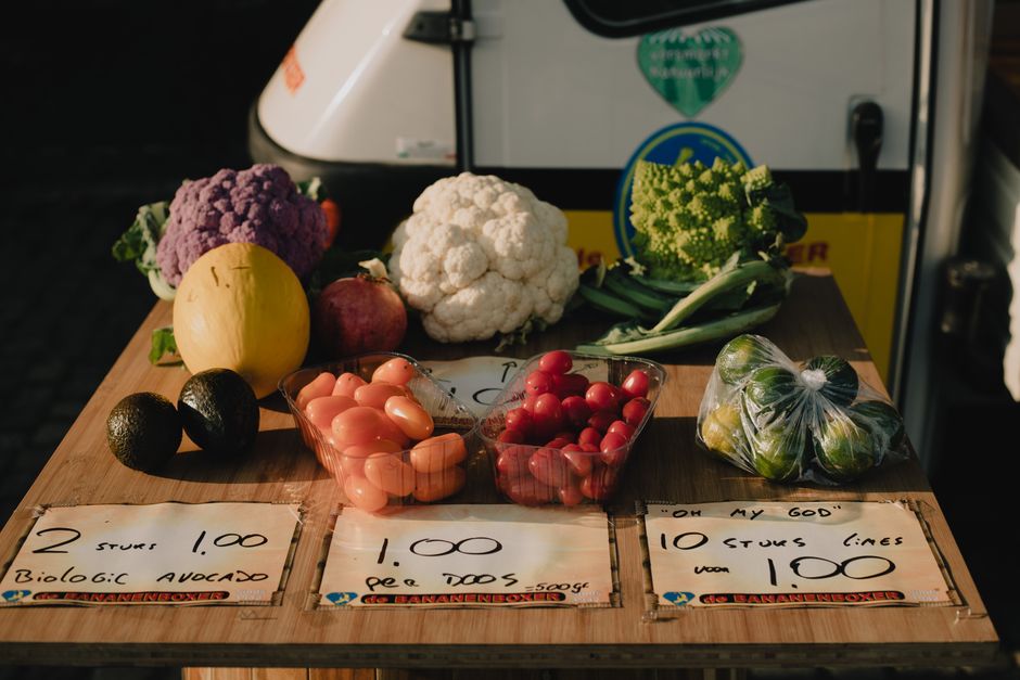 Imagem de uma banca de venda de hortifruti com diversos vegetais, frutas e tomates em um mercado ao ar livre, destacando alimentos naturais e comércio de alimentos frescos.