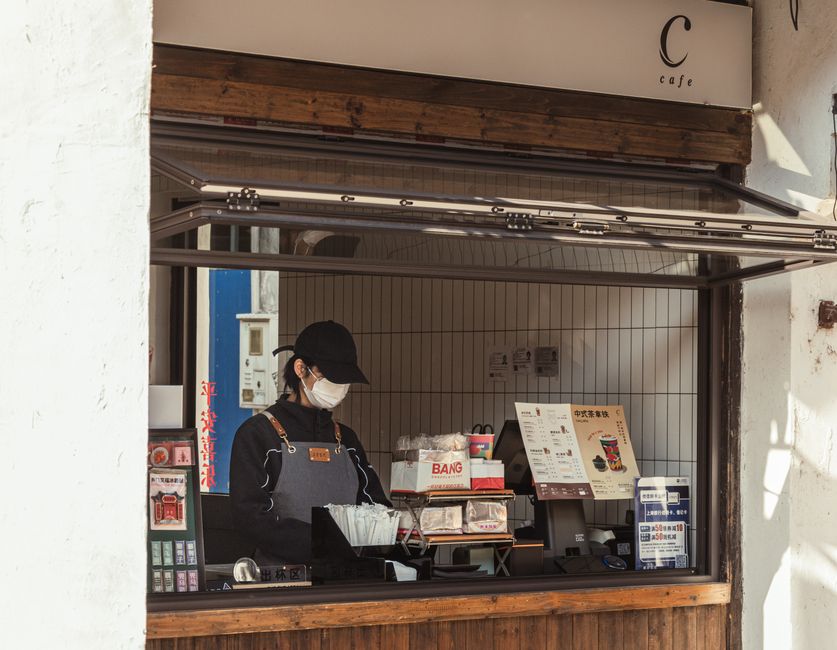 Vendedora atendendo clientes em uma pequena loja de café ou lanchonete.