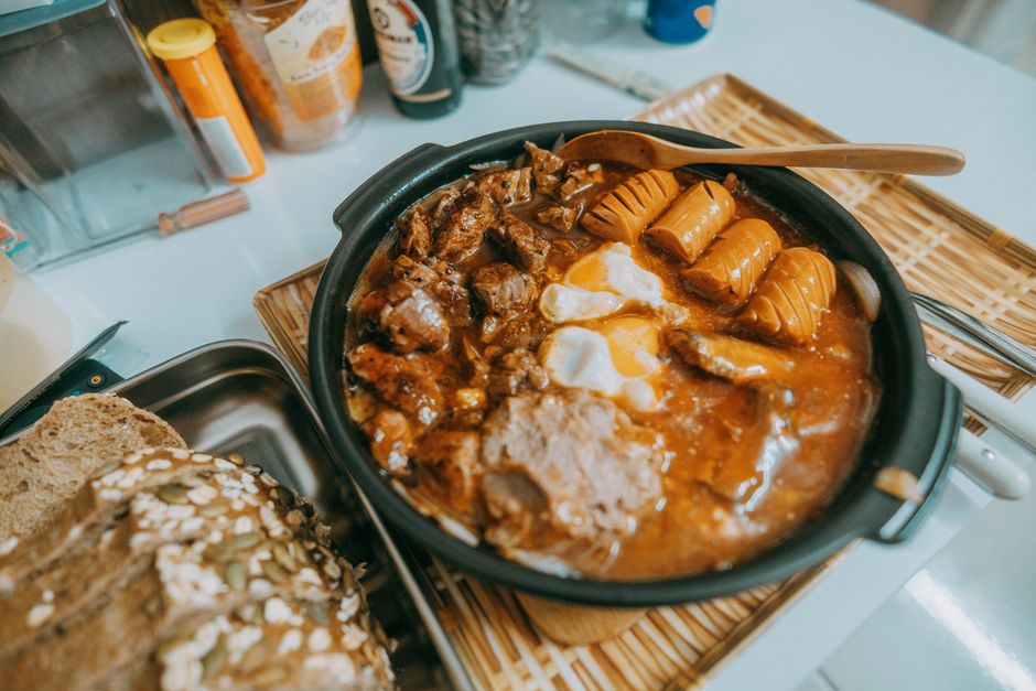 Imagem de uma marmita com comida caseira, incluindo carne, ovos e salsichas, sobre uma mesa de cozinha.