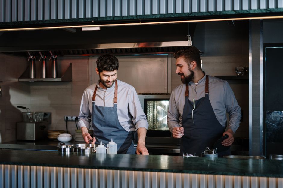 Dois chefs sorrindo enquanto preparam pratos em uma cozinha profissional, simbolizando colaboração e competência culinária.