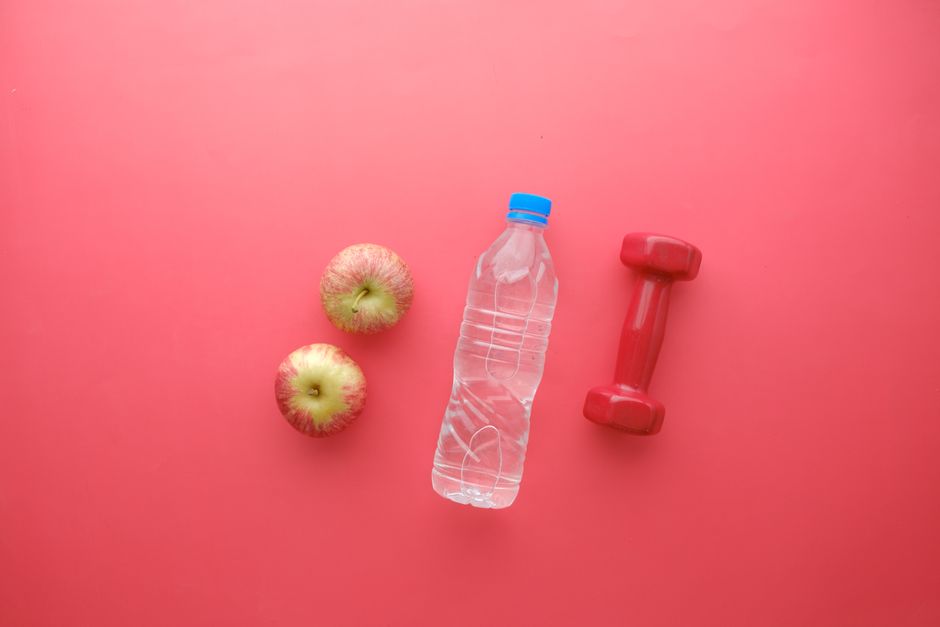 imagem com elementos que representam saúde e bem-estar, como maçãs, água, e um peso de mão, dispostos sobre fundo rosa.