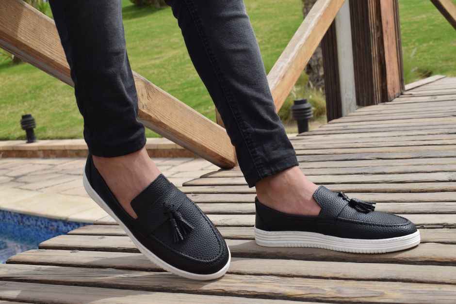Pessoa usando sapatênis preto com detalhe de laço, em ambiente ao ar livre com piso de madeira e jardim ao fundo, transmitindo um estilo casual e confortável.