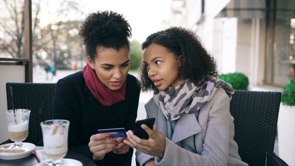 Duas mulheres conversando e olhando uma tela de celular em um ambiente externo, transmitindo conexão e diálogo social.