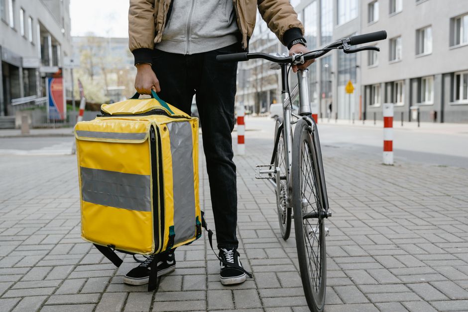 Pessoa com mochila de entrega ao lado de uma bicicleta em ambiente urbano, ilustrando o tema de transportes alternativos e entregas por bicicleta.