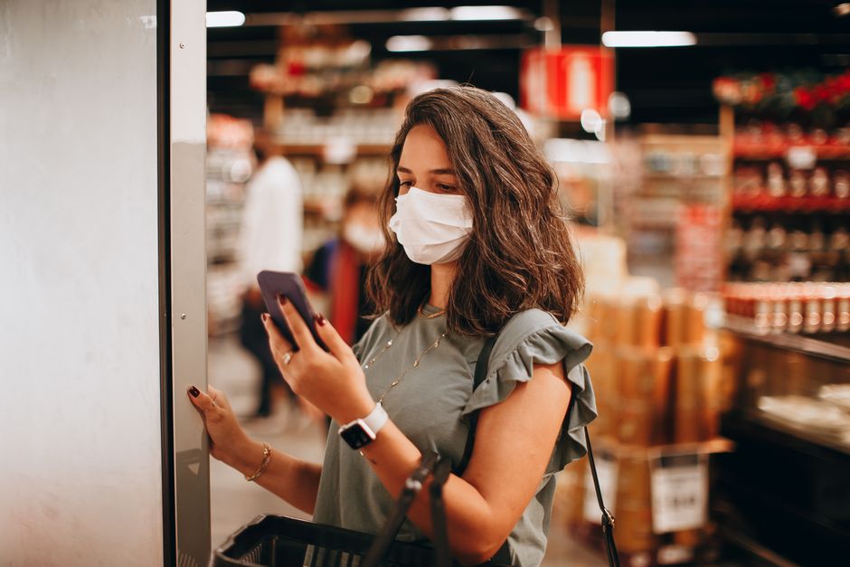 Mulher usando máscara facial enquanto faz compras em supermercado, verificando informações no celular.
