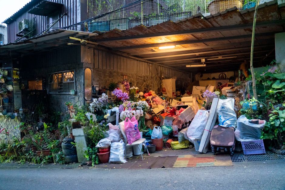 Área de descarte e armazenamento de objetos variados, incluindo flores artificiais, embalagens e móveis, sob uma estrutura de metal e madeira, em ambiente urbano.