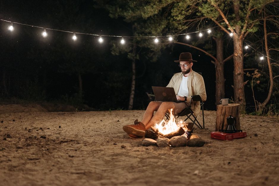 Homem sentado ao ar livre à noite, trabalhando em um computador enquanto desfruta de uma fogueira sob luzes decorativas, em um ambiente natural e tranquilo.
