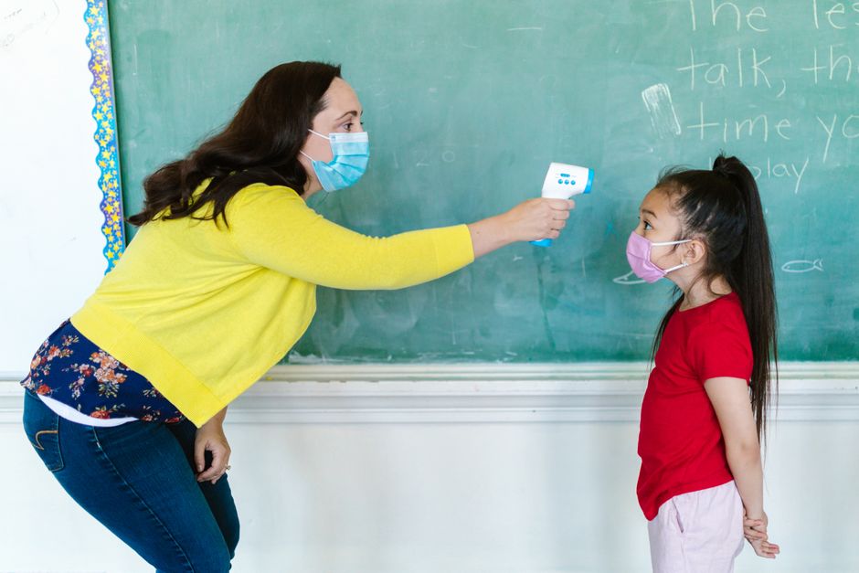 Professora realizando medição de temperatura na criança em sala de aula, simbolizando cuidados com saúde e protocolos durante pandemia.