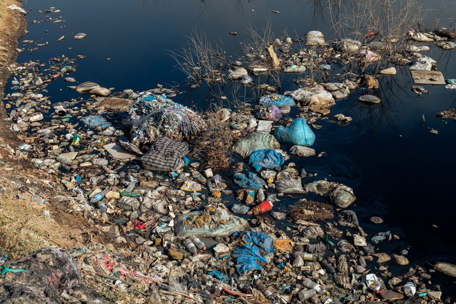 Imagem de um rio poluído com lixo espalhado ao longo da margem, destacando o impacto ambiental da poluição nas águas.