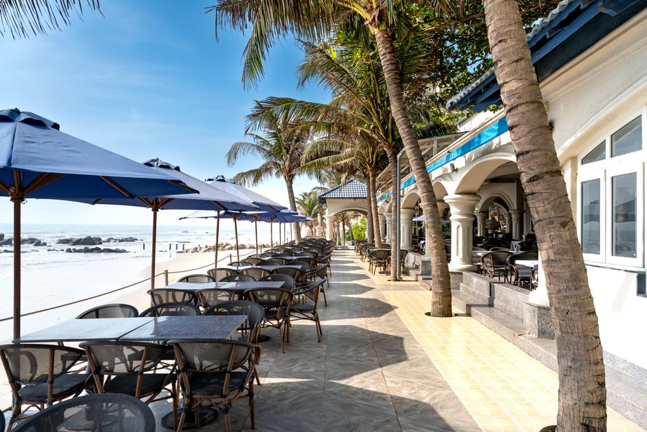 Espaço de restaurante à beira-mar com mesas ao ar livre, sombra de guarda-sóis e palmeiras, ideal para destinos turísticos de lazer.
