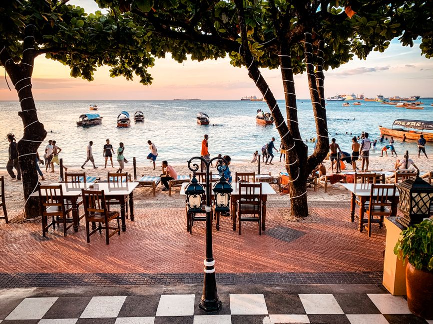 Cenário de uma praia com ambiente de restaurante ao ar livre, mostrando mesas à beira-mar com pessoas aproveitando a vista e o pôr do sol ao fundo.