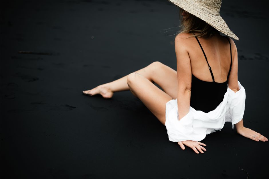 Mulher de chapéu de palha sentada na areia escura, usando roupas de verão e olhando para o mar, transmitindo uma sensação de relaxamento e conexão com a natureza.