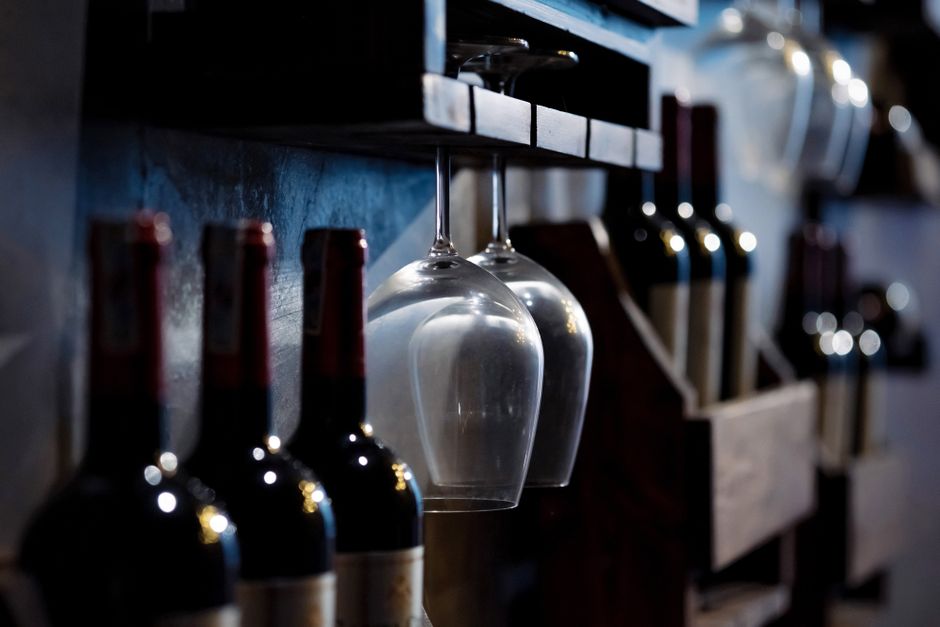 Imagem de uma prateleira de bar com garrafas de vinho e taças penduradas, ideal para ilustrar temas relacionados a gastronomia, bares ou entretenimento social.