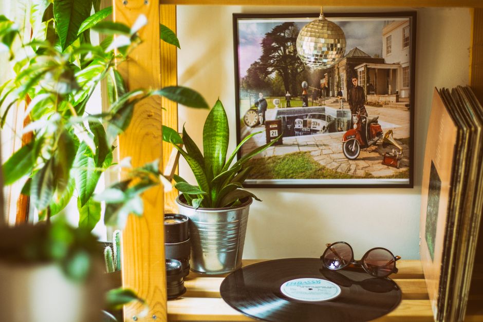 Imagem de um espaço decorado com plantas, uma foto artística na parede, uma vitrola, óculos de sol e discos de vinil, criando um ambiente vintage e aconchegante para blog de decoração ou música.