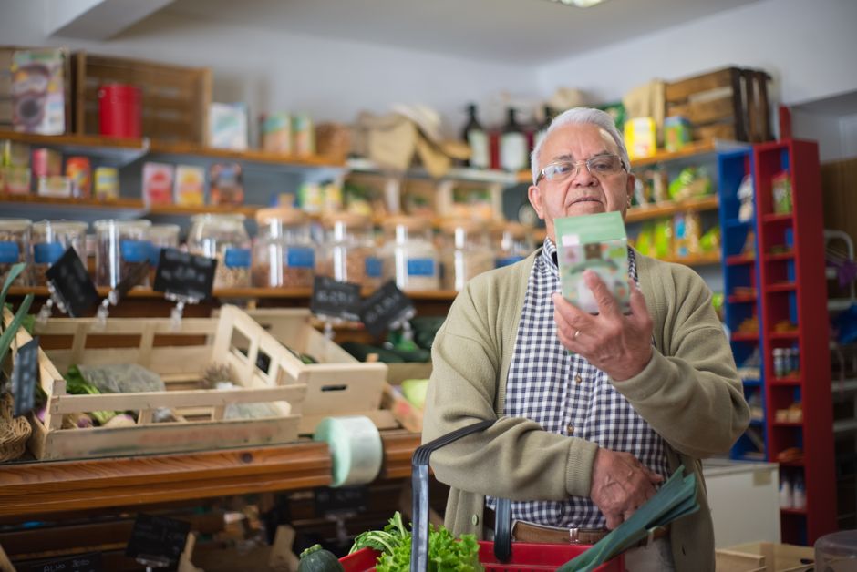 Idoso fazendo compra em mercearia, conferindo uma nota ou dinheiro enquanto escolhe produtos alimentícios.